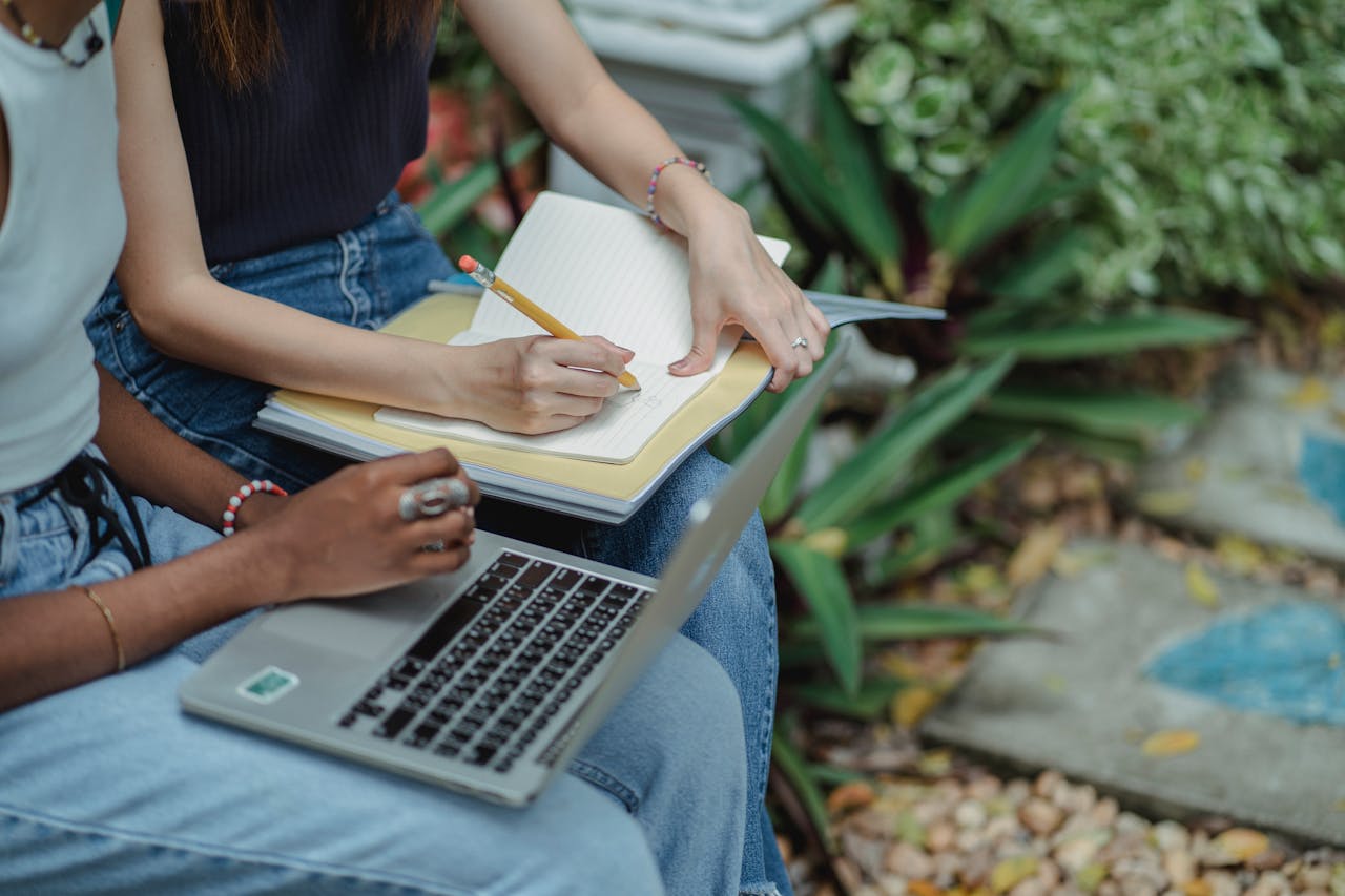 Crop faceless multiracial students in blue jeans using laptop and taking notes in workbook while preparing for exam together in lush summer park